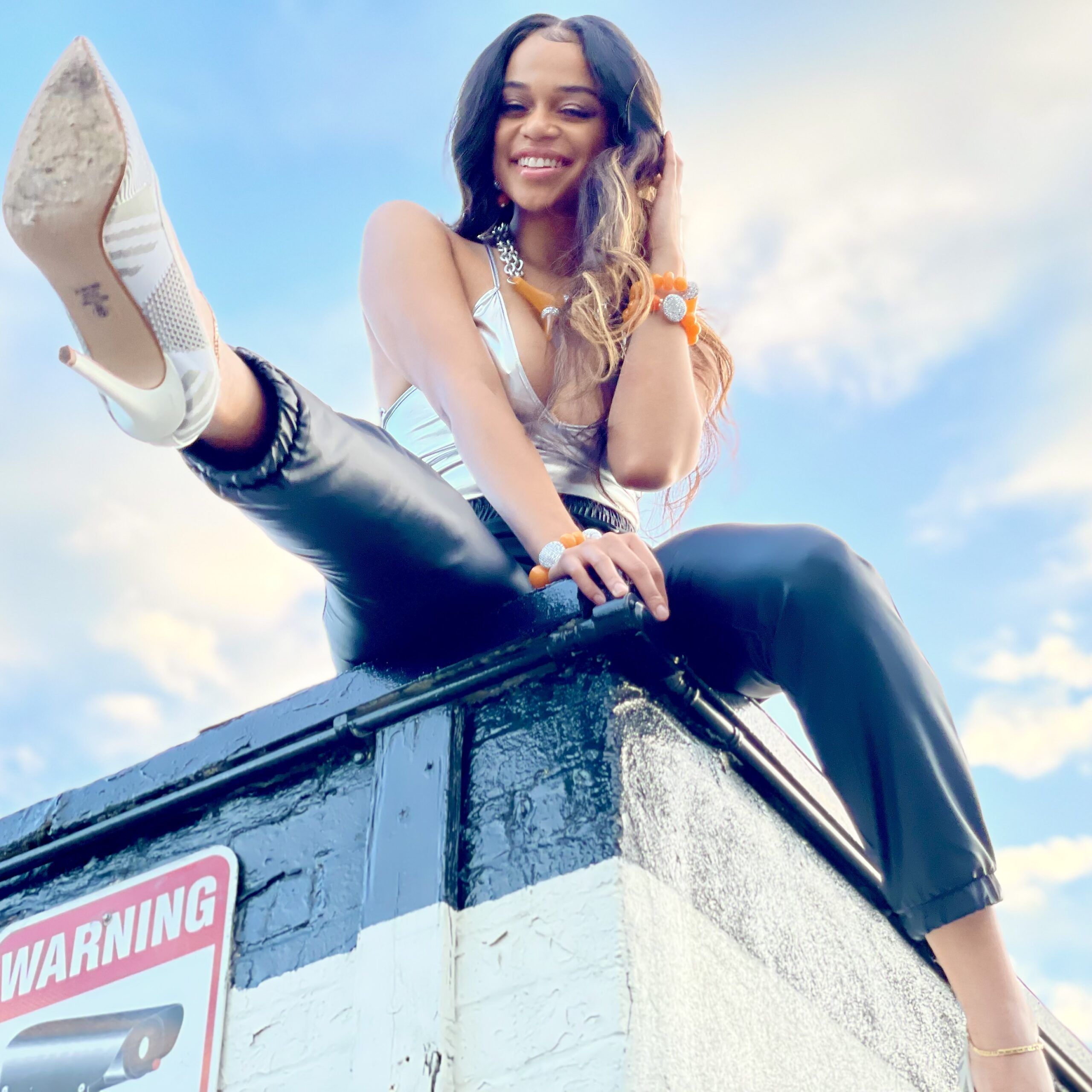 smiling woman sitting on top of a roof looking down wearing a shiny silver tank top and jewelry