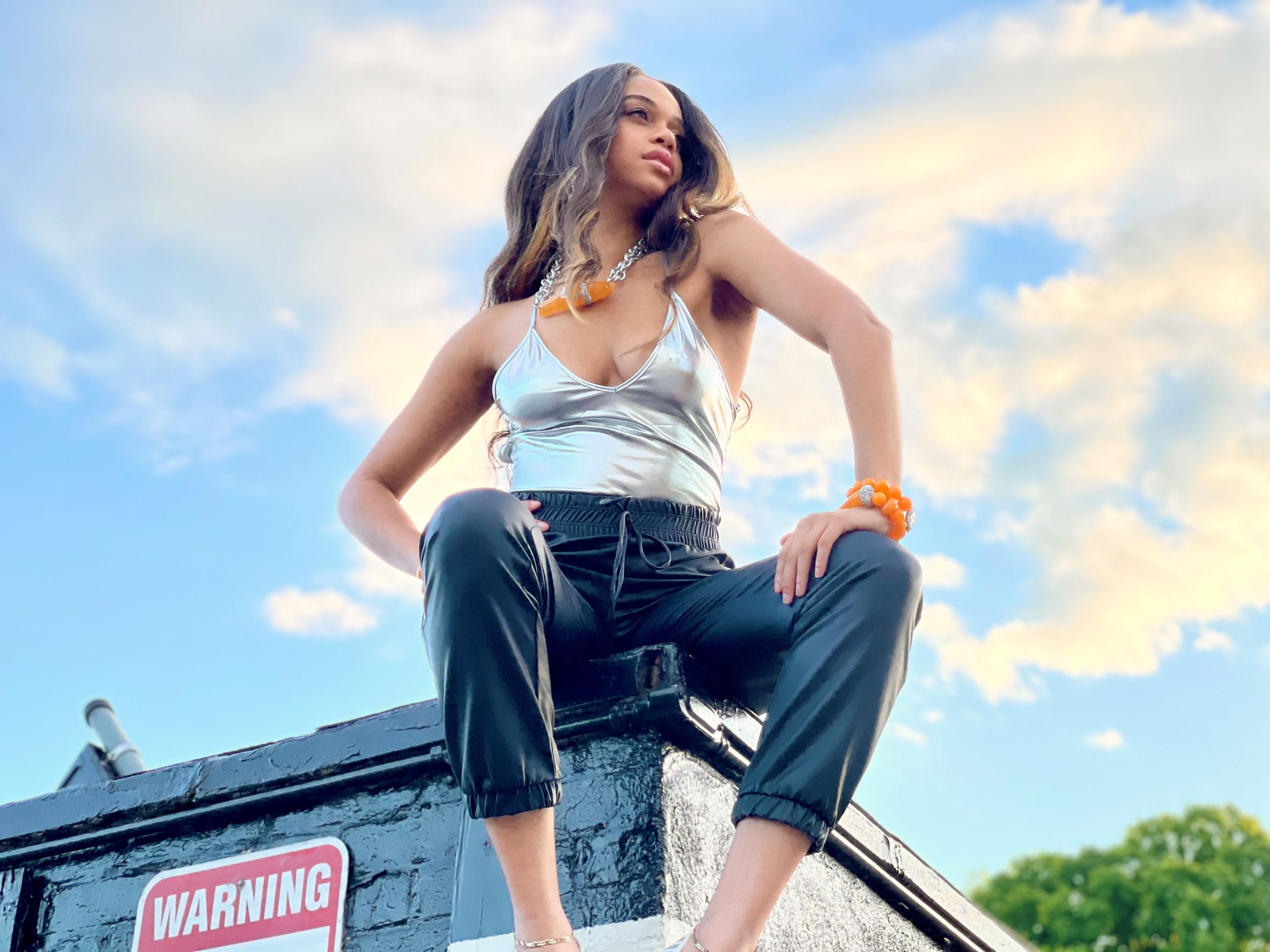 woman sitting on top of a roof wearing a silver tank top wearing chunky orange beaded jewelry with blue sky in background