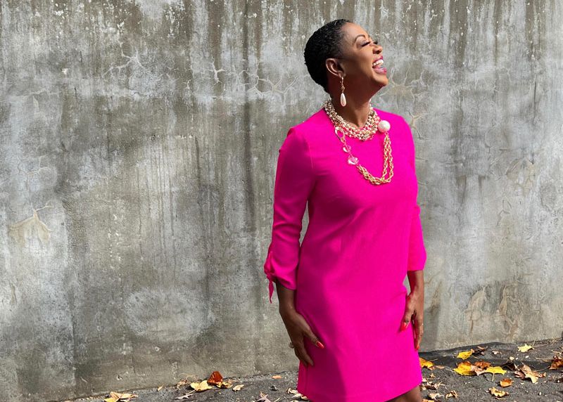 Smiling black woman wearing a bright prink dress looking up to the sky with a cement wall in the background