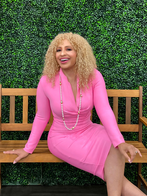 woman with blonde hair wearing a bright pink dress smiling with a long beaded necklace sitting on a park bench