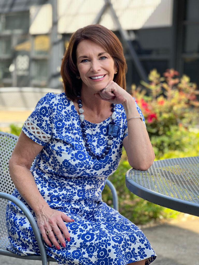 Smiling woman with brown hair wearing a blue and white flowered dress sitting at a table in a garden