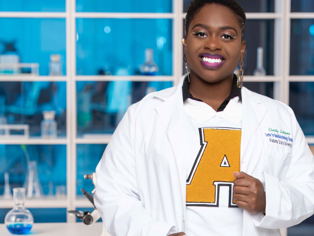 A smiling black woman wearing an Alabama State letterman sweater over a white lab coat standing in a science lab with blue glass accents