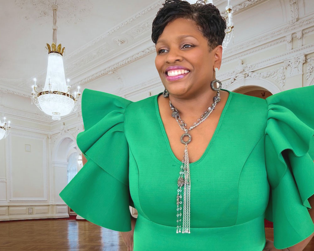 Smiling black woman wearing a bright green dress with large puffy sleeves and a large beaded silver necklace and large hoop earrings standing in a white ballroom with a large chandelier.