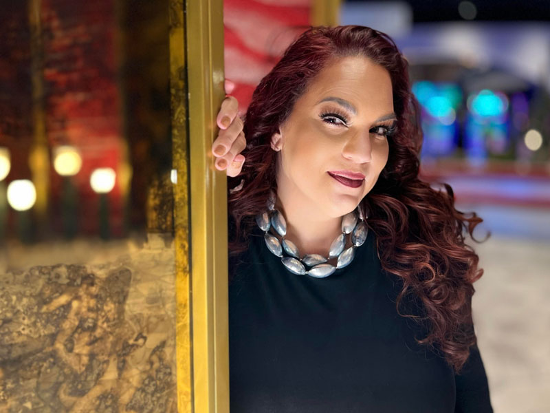 Woman with dark red lipstick and long auburn curly hair wearing a black top with a large silver colored necklace leaning on a gold framed mirror
