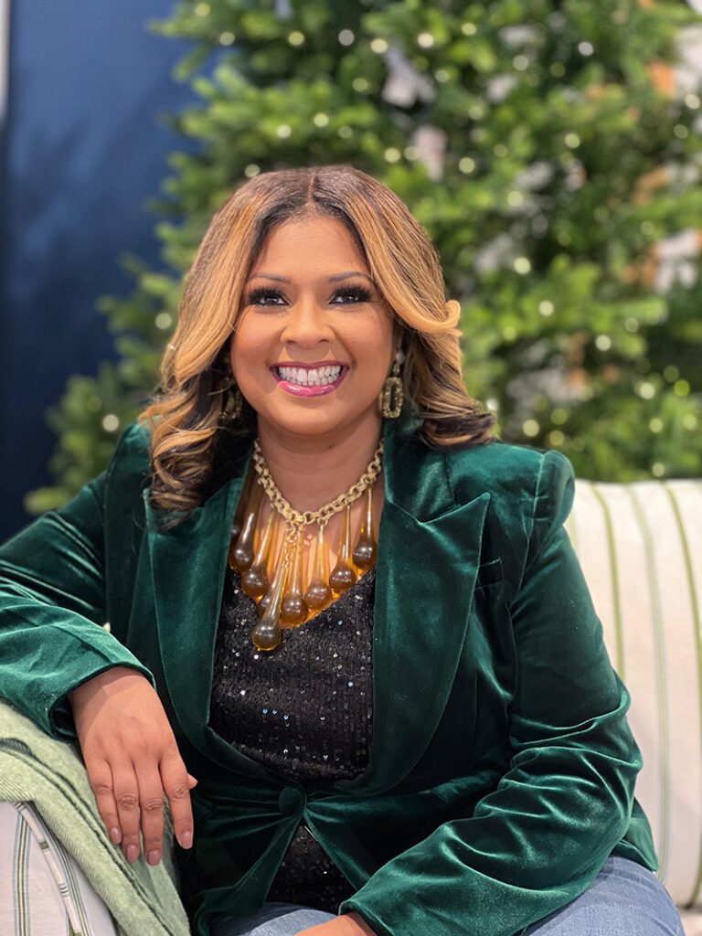 Smiling woman wearing a velvet green jacket sitting in front of a christmas tree and wearing a large necklace with long amber beads