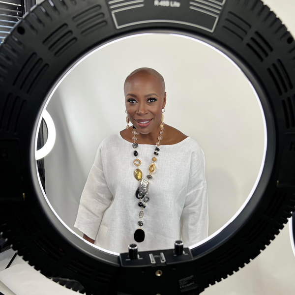 image of smiling black woman staring through a round mirror light wearing a white top and tan pants with a marge beaded necklace in front of a white backdrop