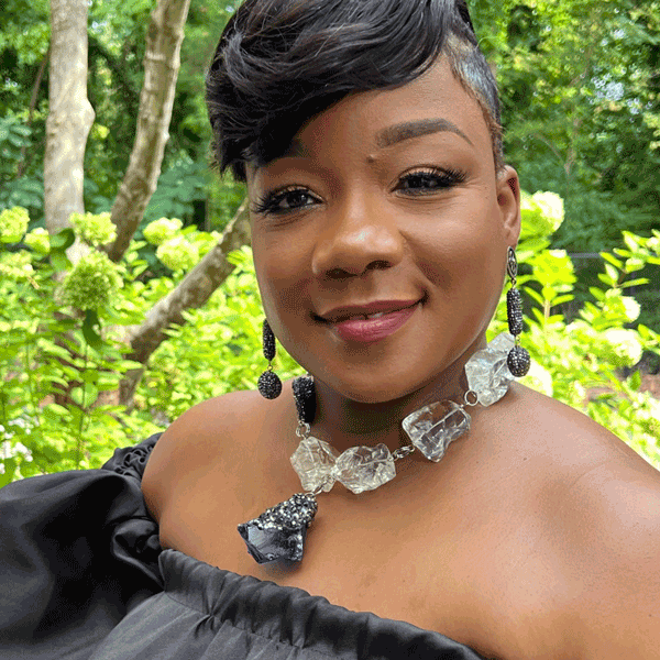 Beautiful black woman wearing a black off the shoulder top and a large beaded necklace and dangling earrings standing in front of greenery.