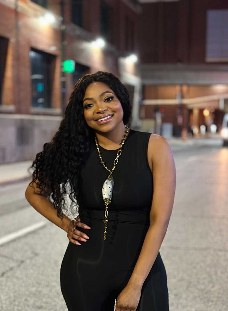 Smiling black woman standing in front of a night time cityscape wearing a black sleeveless jumpsuit and and a long lariat style necklace with a large bead element