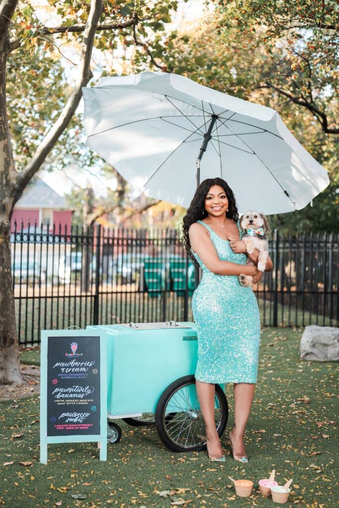 Image of a smiling black woman wearing a turquoise sequined sleeveless dress standing in front a a turquoise ice cream cart holding a small white dog in a part setting with a white umbrella